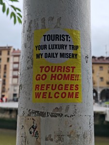 on a whitewashed pole, a sticker that reads: "Tourist: your luxury trip my daily misery" and "Tourist go home!!! Refugees welcome"